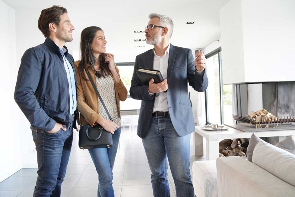 A couple discusses paperwork at a dining table with a house for sale sign visible through a window.